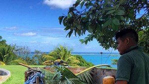A man peeling a coconut on a wooden table outdoors with a scenic ocean view in the background.