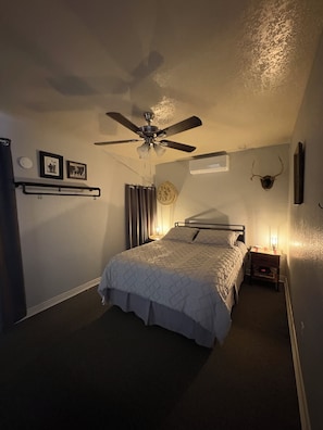 A bedroom with a bed, a ceiling fan, a nightstand with a lamp, and a wall-mounted picture frame.