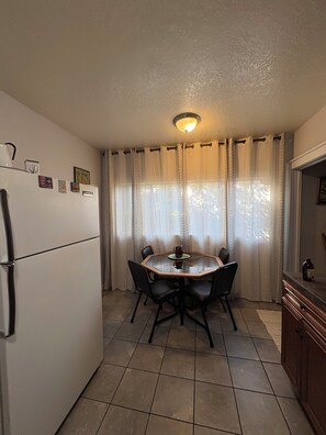 A compact kitchen with a dining table, chairs, a refrigerator, and a window with curtains.