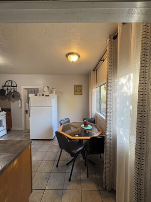 Une cuisine compacte équipée d’un réfrigérateur, d’une cuisinière et de casseroles suspendues. Une table à manger avec des chaises est placée près d’une fenêtre ornée de rideaux à motifs.