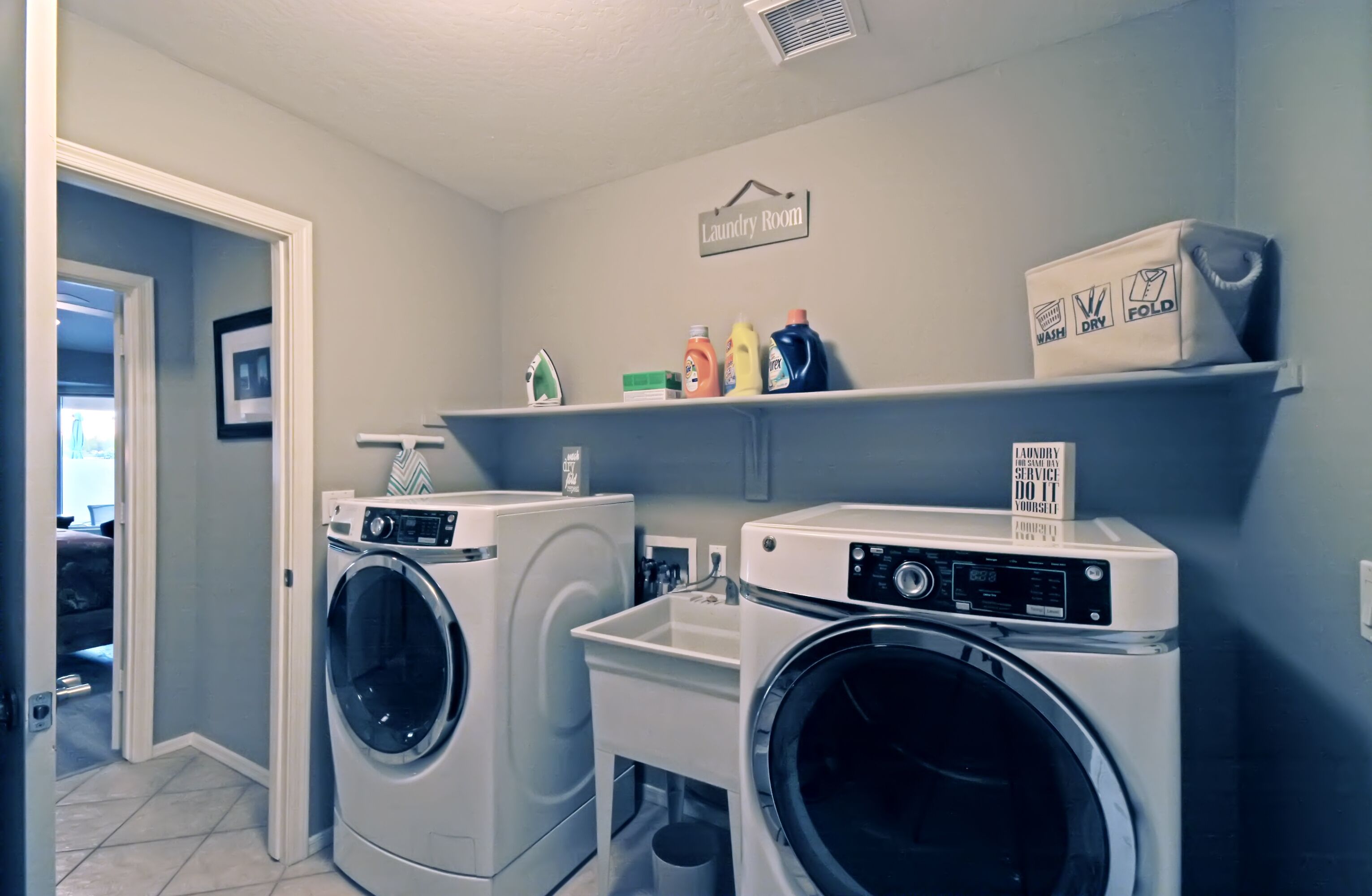 A laundry room with two front-loading washing machines, a shelf with laundry supplies, and a sign indicating it's a self-service area.