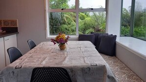 Un coin repas avec une table recouverte d’une nappe à motifs de journal, un vase de fleurs et un banc agrémenté de coussins.