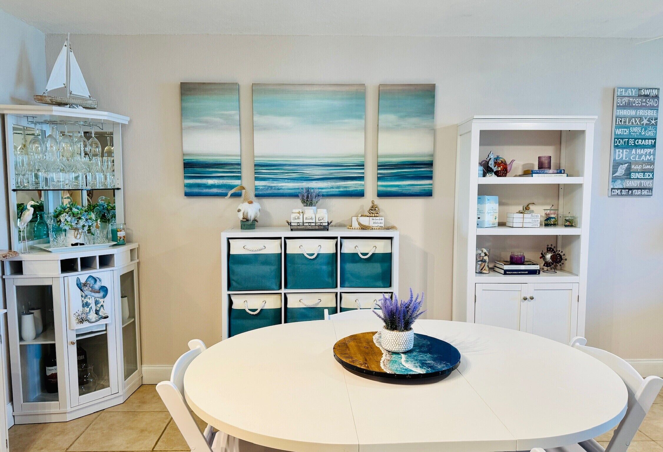 A dining area with a round table, white cabinets, and a wall with three ocean-themed paintings.
