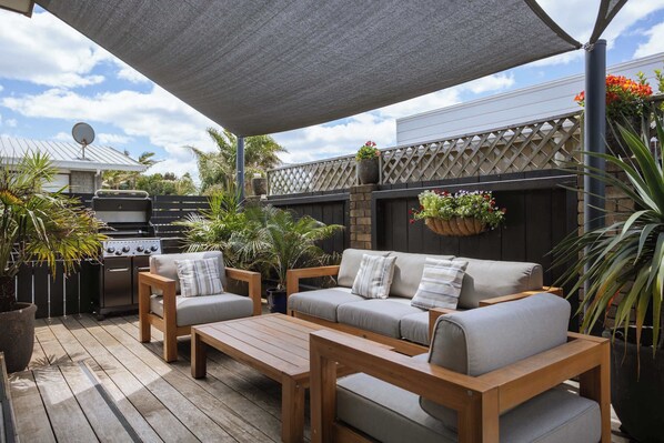 An outdoor patio area with a wooden deck, a barbecue grill, and a seating arrangement including a sofa and two chairs.