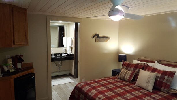 Ein Schlafzimmer mit Bett, Nachttisch, Lampe, Deckenventilator und angrenzendem Badezimmer.