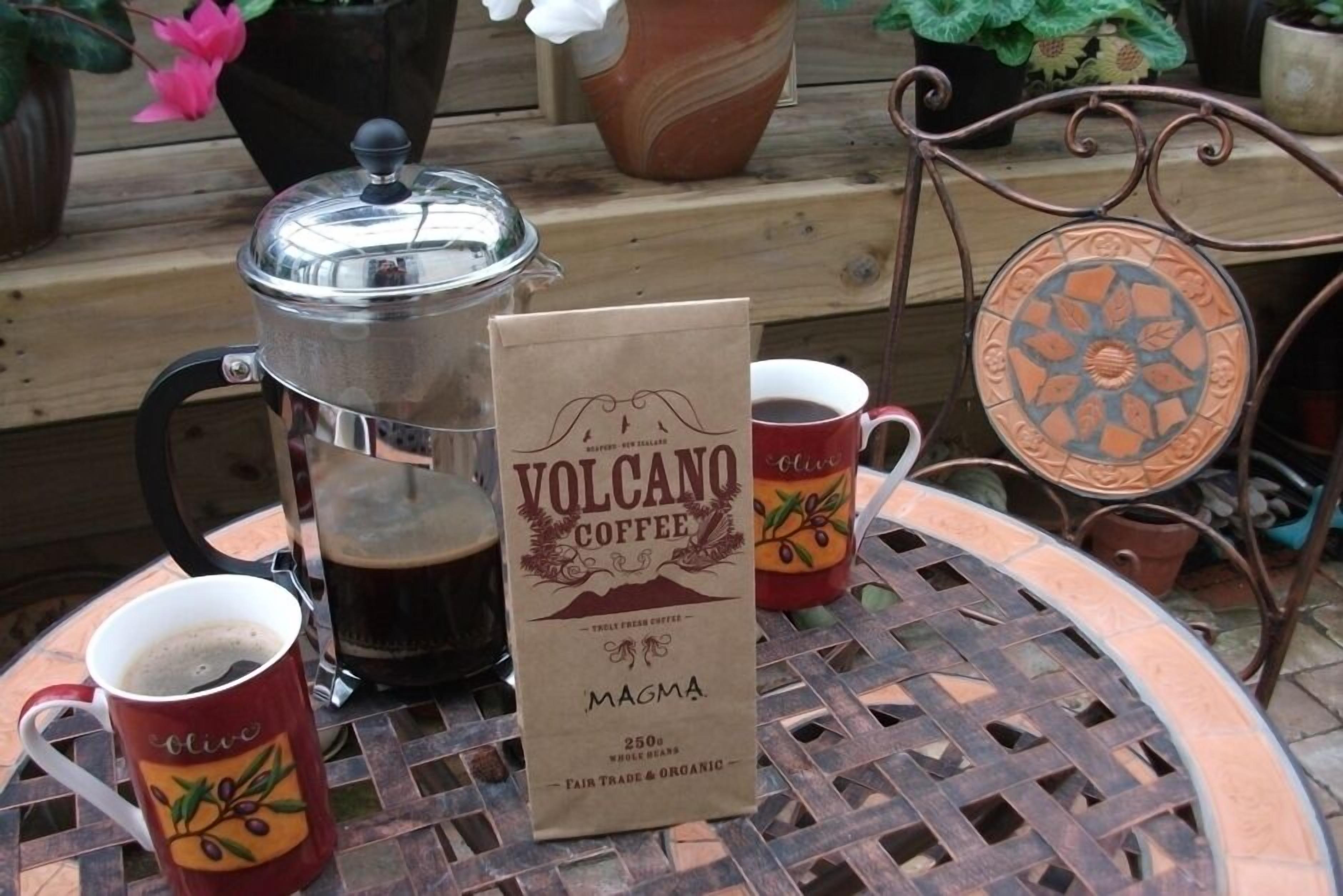 Koffie-opstelling met een French press, twee mokken, een papieren zakje met de tekst 'Volcano Coffee Magma' en een decoratief bord op een ronde metalen tafel.