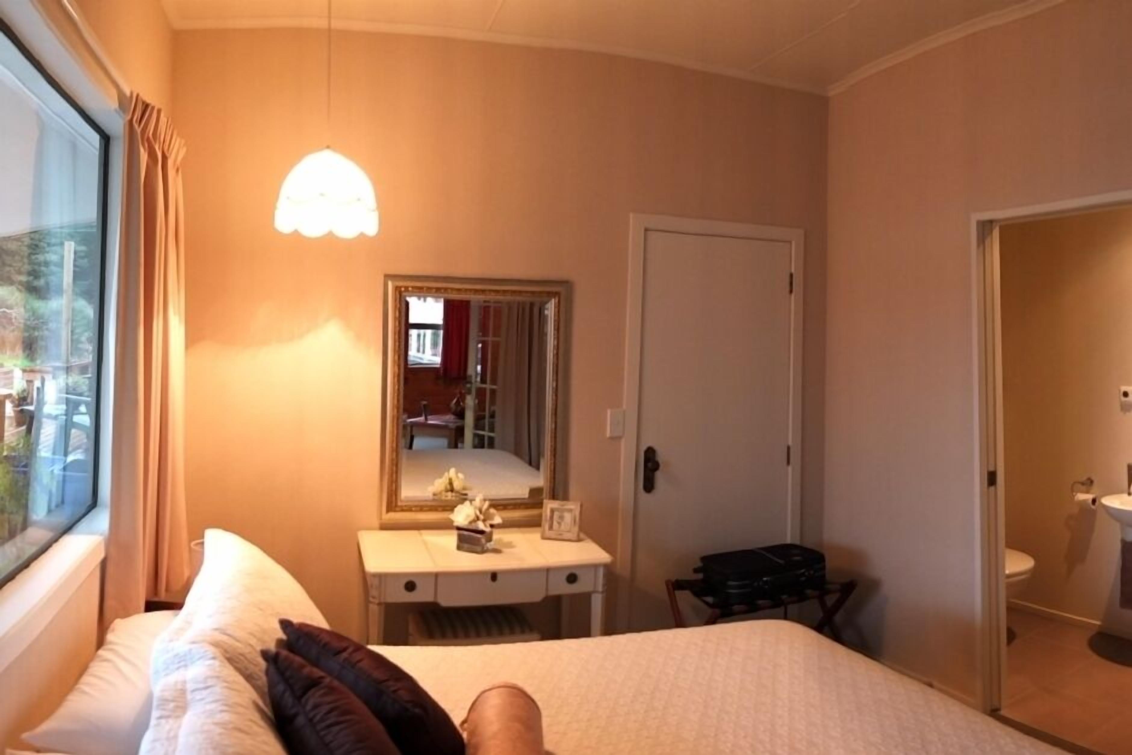 A hotel room with a bed, a desk, a mirror, a window with curtains, and a door leading to a bathroom.
