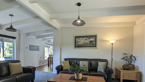 Ein Wohnzimmer mit einem dunkelbraunen Ledersofa, einem Holztisch und einer Stehlampe. An der Wand hängt ein gerahmtes Bild und durch das Fenster ist viel Grünlandschaft zu sehen.