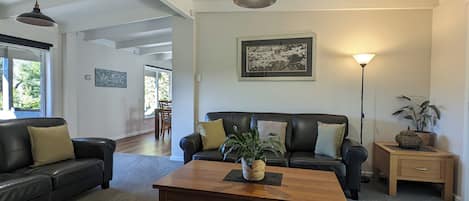 Sala de estar con un sofá de cuero oscuro, una mesa de centro de madera y una lámpara de pie. Se observa un cuadro en la pared y una ventana con vistas a la vegetación.