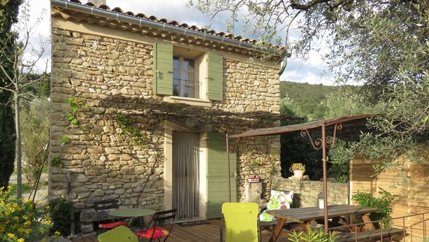 A stone house with a wooden deck, a green chair, a table, and a pergola.