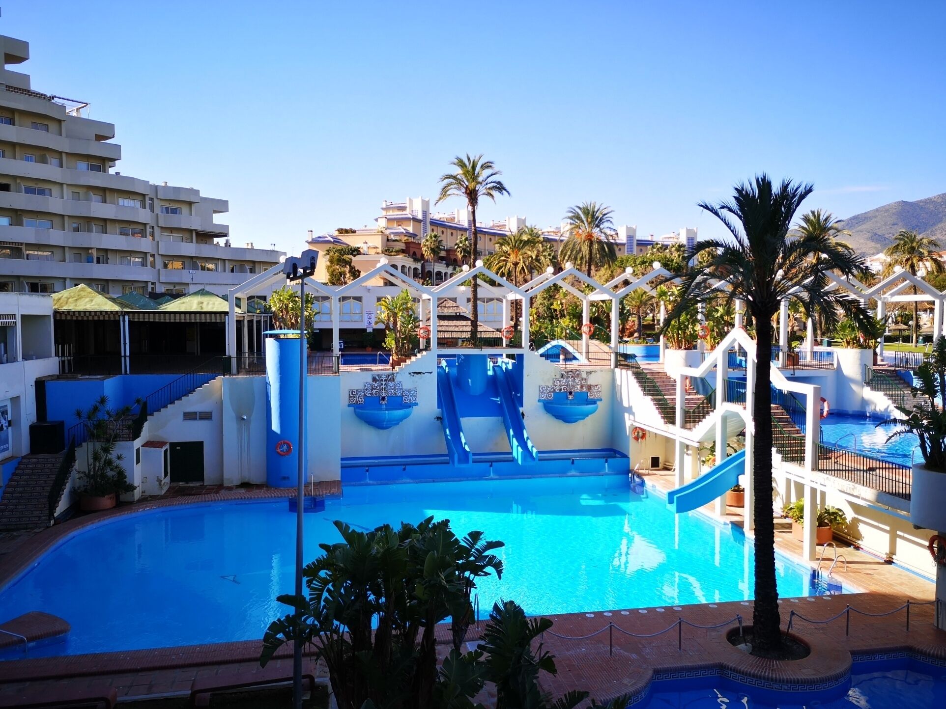 Una piscina con toboganes, palmeras y vistas a edificios de hotel al fondo.