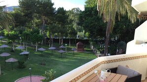 A balcony with a wooden table and a wicker chair, overlooking a well-maintained garden with grass, trees, and thatched umbrellas. - Beautiful apartment on the beach. A Luxury. (Benalmádena)