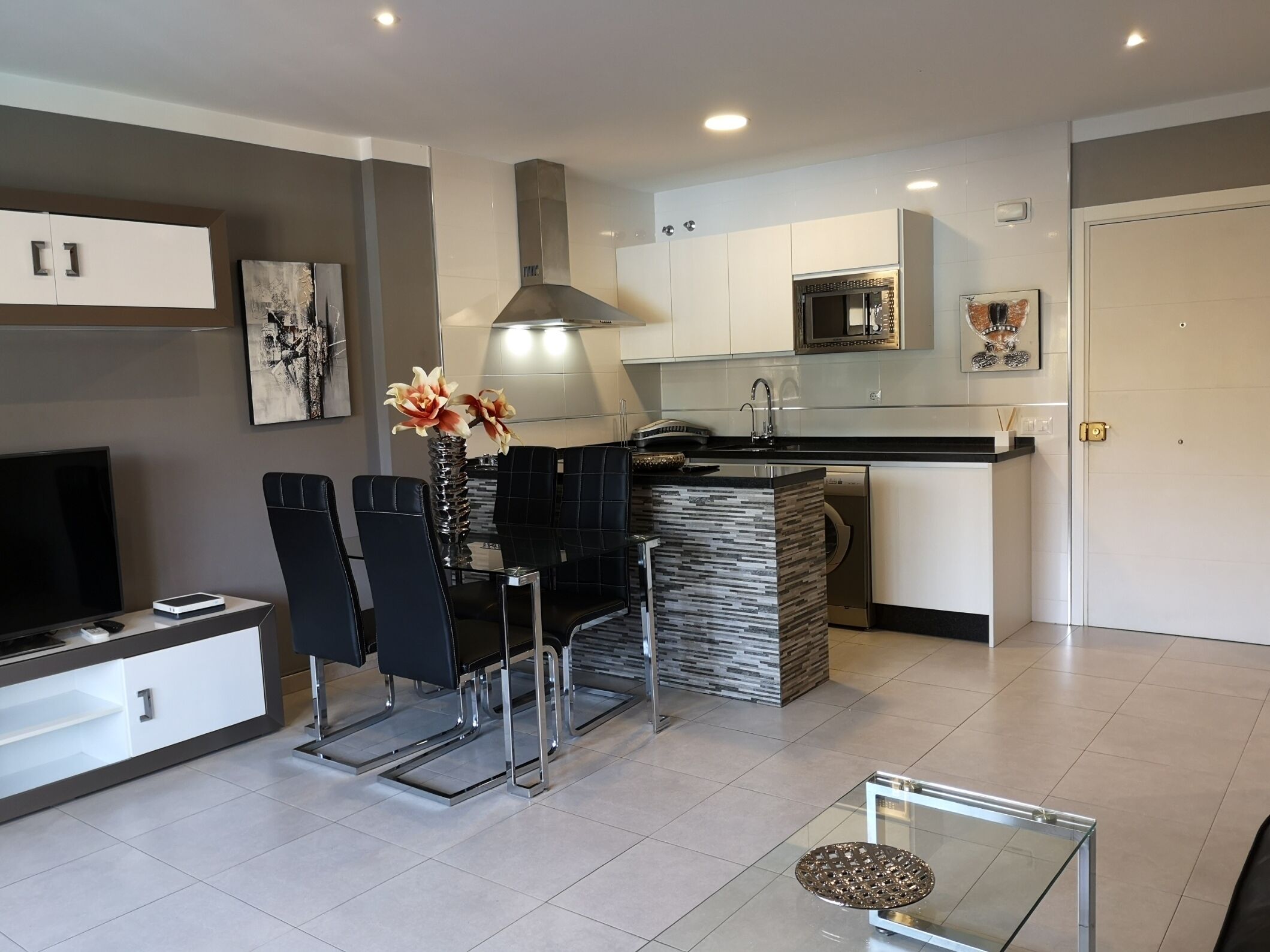 Een moderne keuken met een eethoek, met een zwarte eethoekset, een glazen salontafel en een televisie op een meubel.