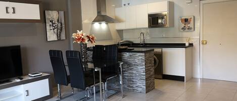 Een moderne keuken met een eethoek, met een zwarte eethoekset, een glazen salontafel en een televisie op een meubel.