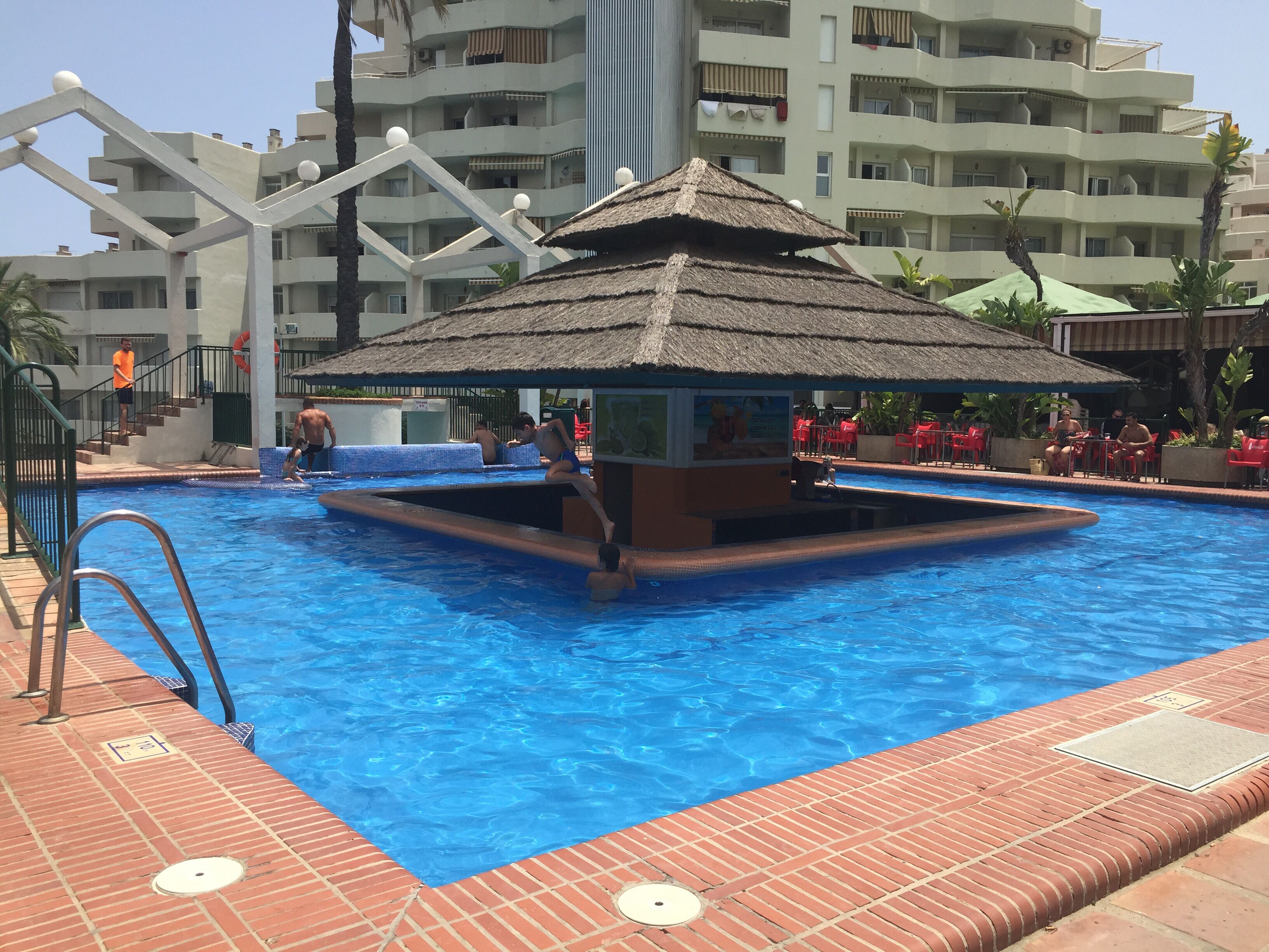 Ein Schwimmbecken mit einer strohgedeckten Struktur, umgeben von einer Ziegelterrasse und roten Stühlen.