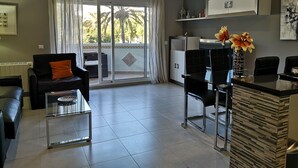 A modern living room with a sofa, a glass coffee table, a dining table with chairs, a kitchen area, and a sliding glass door leading to a balcony with a view of palm trees. - Beautiful apartment on the beach. A Luxury. (Benalmádena)