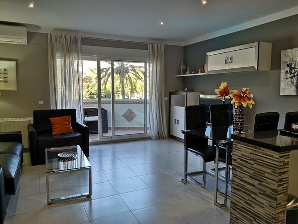 A modern living room with a sofa, a glass coffee table, a dining table with chairs, a kitchen area, and a sliding glass door leading to a balcony with a view of palm trees. - Beautiful apartment on the beach. A Luxury. (Benalmádena)