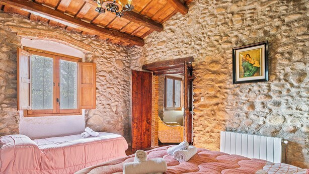 Un dormitorio rústico con paredes de piedra, vigas de madera, una cama con ropa de cama rosa y una ventana con contraventanas de madera.