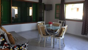 A dining area with a table and chairs, a sofa, a television, and large windows with green shutters. - Casangeles Holiday Homes (Mozaga)