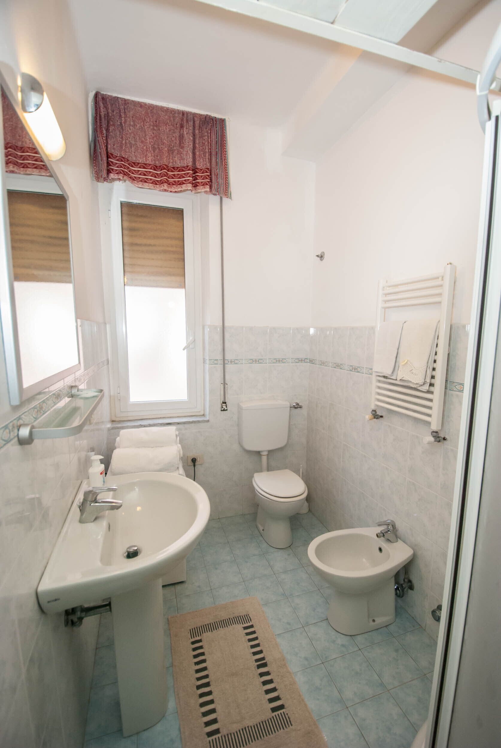 A bathroom with a sink, toilet, bidet, and a window with a curtain.