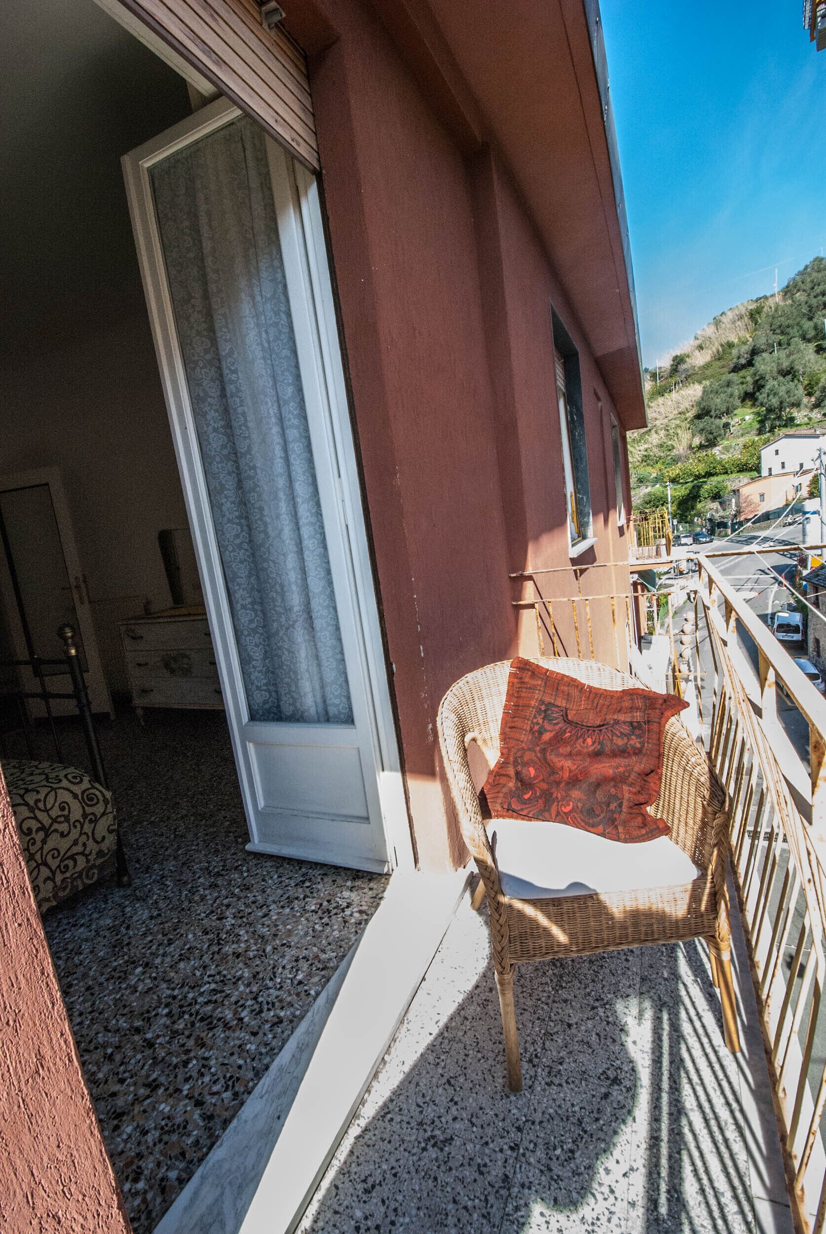 Ein Balkon mit Korbstuhl, rotem Kissen, einer Schiebeglastür und Blick auf eine Straße und Hügel.