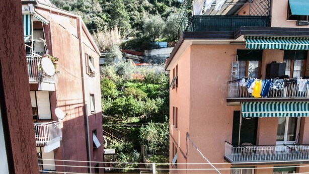 Un balcon avec un fauteuil en osier, une rambarde en métal et une vue sur les bâtiments et les arbres voisins.