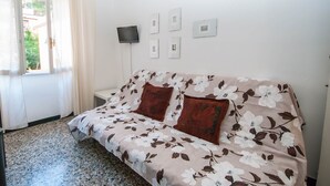 Une pièce avec un canapé à motifs floraux, un téléviseur à écran plat fixé au mur et une fenêtre avec des rideaux.