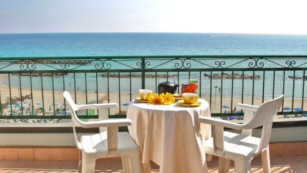 Een balkon met een tafel voor twee, uitkijkend op een strand met parasols en ligstoelen.