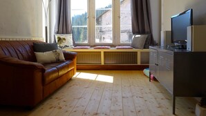 Una sala de estar acogedora con un sofá de cuero marrón, un rincón con cojines junto a la ventana, un televisor sobre un mueble bajo y un radiador debajo de la ventana.