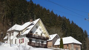 Una casa cubierta de nieve con techo inclinado, un balcón y un edificio de piedra cercano.