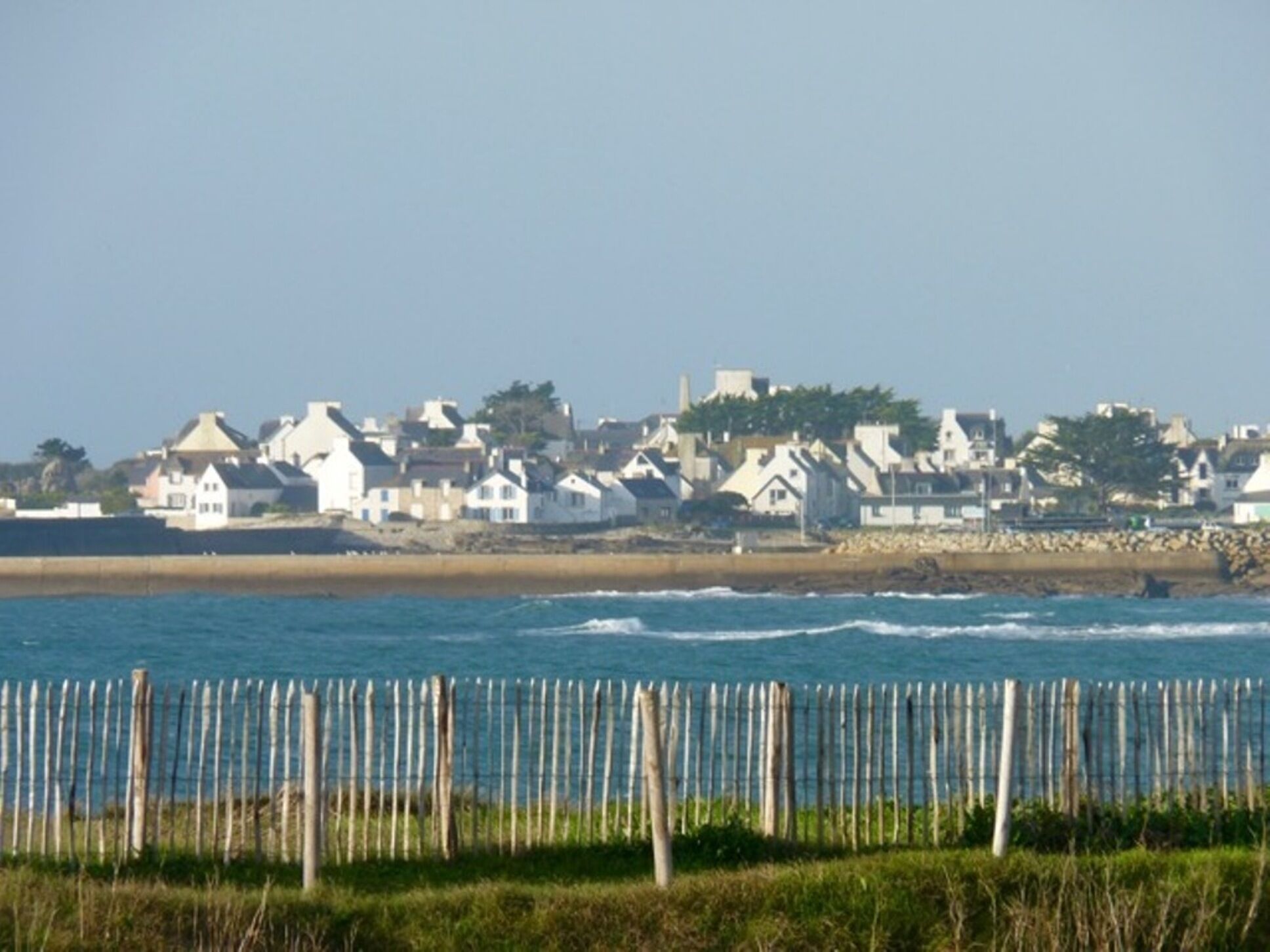 Een kustplaatsje met huizen aan het strand, een houten hek op de voorgrond en de zee op de achtergrond.