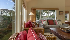 Amplia sala de estar con un gran ventanal que ofrece vistas a palmeras y casas. Hay un sofá de mimbre con cojines rojos y un sofá gris con almohadones estampados. Una mesa de centro de madera está en primer plano.