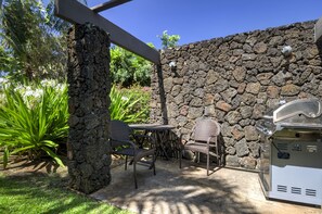 Eine Terrasse im Freien mit einer Steinmauer, zwei Korbstühlen, einem Grill und üppigem Grün.