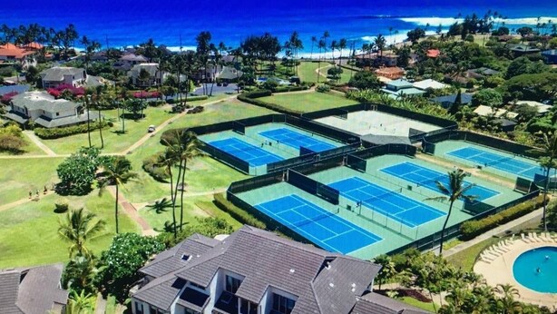 Complejo de canchas de tenis con varias pistas, rodeado de edificios residenciales y palmeras, con vistas a una playa.