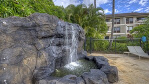 Una cascada de roca con una piscina, rodeada de exuberante vegetación y palmeras, con un edificio al fondo.