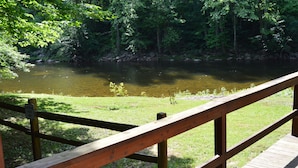 A wooden deck overlooking a river with a grassy area and trees in the background. - Stay WATERFRONT. Stay CHEAT RIVER LODGE (Elkins)