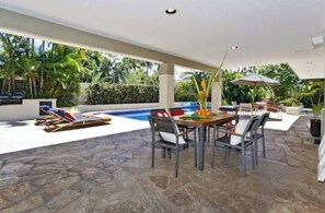 En udendørs patio med pool, stenbelægning, et spisebord med stole og et loungeområde med liggestole.