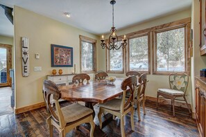 Una sala da pranzo con tavolo e sedie in legno, ampie finestre vista alberi, un lampadario e un orologio appeso al muro.