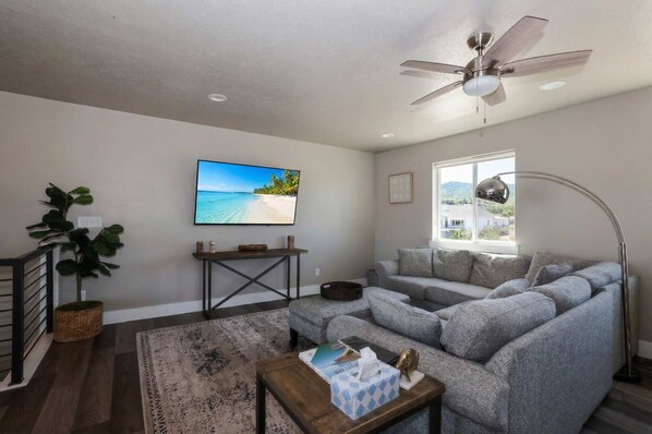 A living room with a grey sofa, a television, a ceiling fan, a window with a view, and a plant.