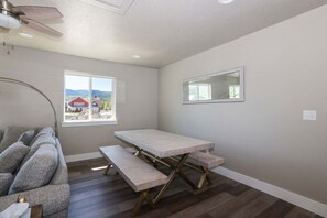A room with a large window, a ceiling fan, a table with benches, and a view of a red house.