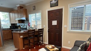 A modern kitchen with wooden cabinets, a central island, and a dining area with chairs. A dark wooden door with a sign is visible, and a living area with a black leather sofa and a patterned rug is in the foreground. - High end 1BR/1BA Attached House in the Heart of Silicon Valley, Non-Smokers only (San Jose)