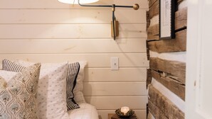 A bedroom with a bed, a bedside table with a candle, a window with a lamp, and wooden paneling. - Modern Cabin By Bear Lake (Cookeville)