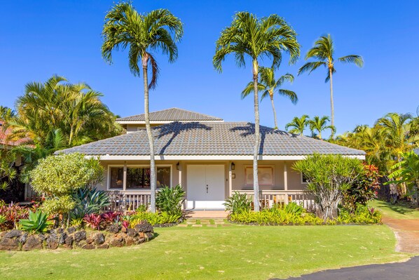 Une maison à deux étages, avec un toit recouvert de tuiles, entourée d’une végétation tropicale et de palmiers.