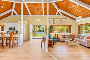 A spacious living area with a high ceiling, a kitchen with white cabinets, and a view of a green landscape through large windows.