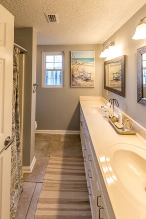 Une salle de bain avec un long lavabo blanc, une fenêtre et un tableau représentant une scène de plage.