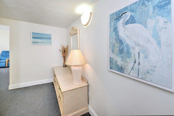 A room with a wooden dresser, a lamp, a framed painting of a bird, and a wall-mounted mirror.