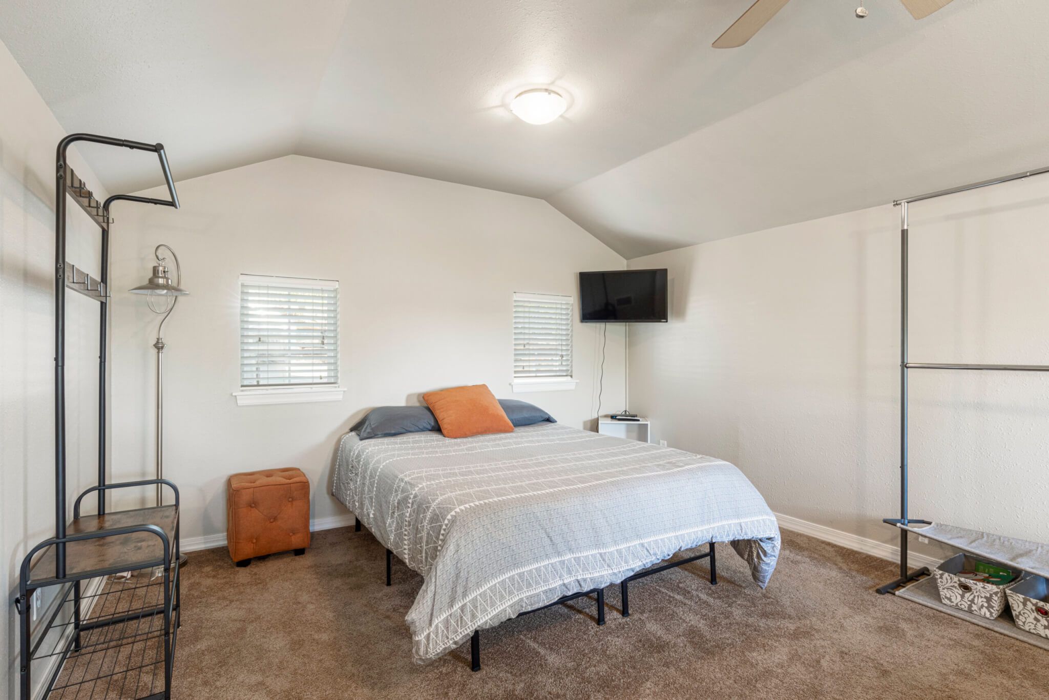 A bedroom with a bed, a TV mounted on the wall, a ceiling fan, and a window with blinds.