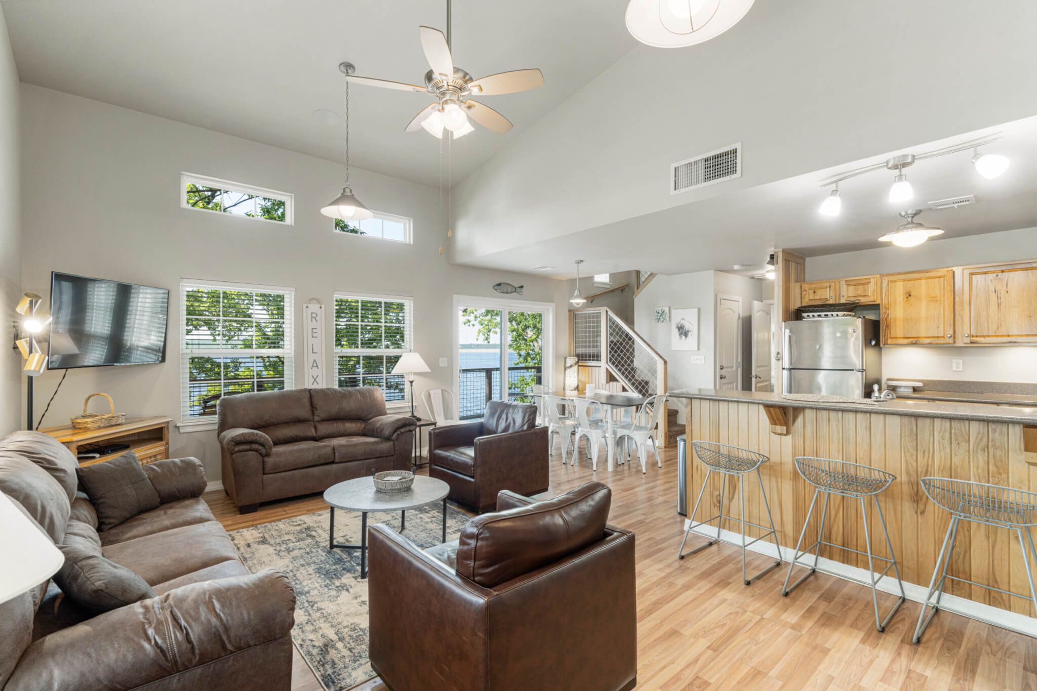 A spacious living area with a large sofa set, a coffee table, and a dining area with bar stools. There is a kitchen with modern appliances and wooden cabinets.