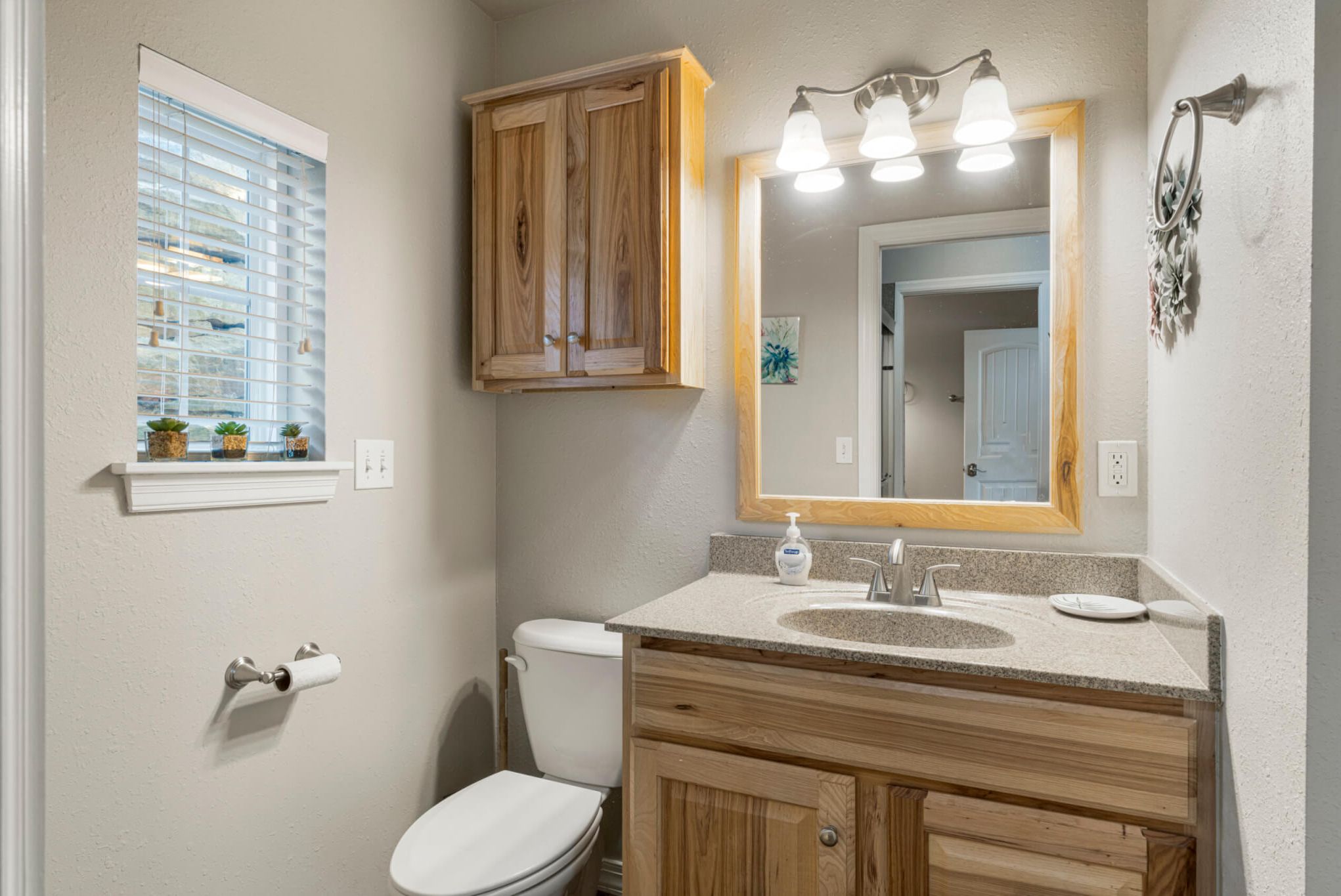 A bathroom with a toilet, sink, wooden cabinets, a mirror with lights, and a window with blinds.