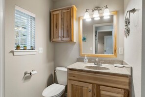 A bathroom with a toilet, sink, wooden cabinets, a mirror with lights, and a window with blinds. - Hideaway at CrossTimbers Marina (Sperry)